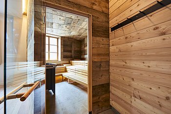 Die Glastür der Sauna aus Holz steht offen und wir können einen Blick auf die leeren Bänke darin werfen. Ein Fenster gegenüber lässt Licht herein.