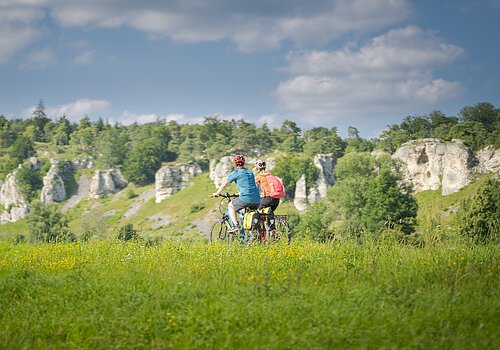Zwei Personen fahren mit Fahrrädern auf einem grasbewachsenen Feld vor einer Felslandschaft.