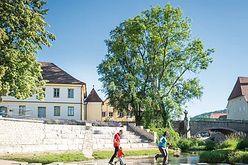 Zwei Personen überqueren auf Trittsteinen einen flachen Fluss, im Hintergrund Gebäude und Bäume.
