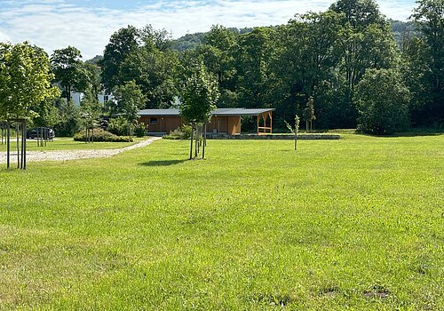 Grill- und Zeltplatz Berching Grüne Wiese mit jungen Bäumen, im Hintergrund ein Holzgebäude und Wald. Ein Kiesweg führt zum Gebäude.