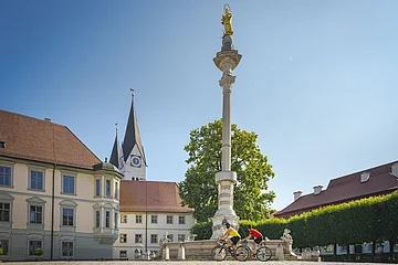 Zwei Radfahrer fahren an einer Mariensäule auf einem Platz mit historischen Gebäuden vorbei.