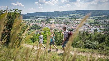 Drei Personen wandern auf einem Pfad mit Blick auf eine Stadt und bewaldete Hügel im Hintergrund.