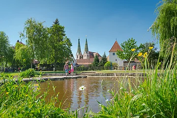Park mit Teich, Wiese, Bäumen und Menschen vor historischen Gebäuden mit zwei Türmen bei blauem Himmel