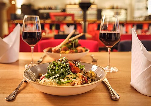 Tisch mit zwei Tellern, zwei Gläsern Rotwein, Besteck und zwei gefalteten weißen Servietten in einem Restaurant.