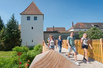 Fünf Personen mit Rucksäcken gehen auf einem gepflasterten Weg neben einem weißen Turm mit rotem Dach bei klarem Himmel.