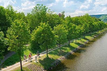 Fünf Radfahrer auf einem Weg entlang eines Flusses mit Bäumen und Hügeln im Hintergrund bei bewölktem Himmel.