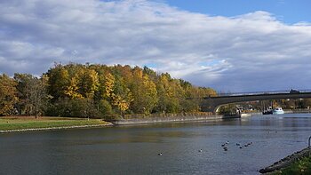Fluss mit Enten, Brücke und herbstlich gefärbten Bäumen unter bewölktem Himmel.