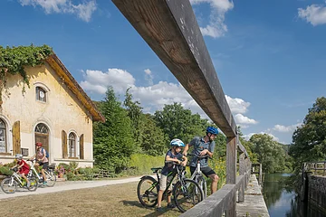 Familie mit vier Personen und Fahrrädern neben einem alten Haus und einem Fluss unter blauem Himmel.